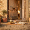 A gold pineapple ornament, a loaf of bread, a small dish of salt, and a ribbon-wrapped gift arranged on a doormat at a warm, sunlit front door strung with string lights and flanked by potted olive trees — traditional housewarming gifts on a doorstep.