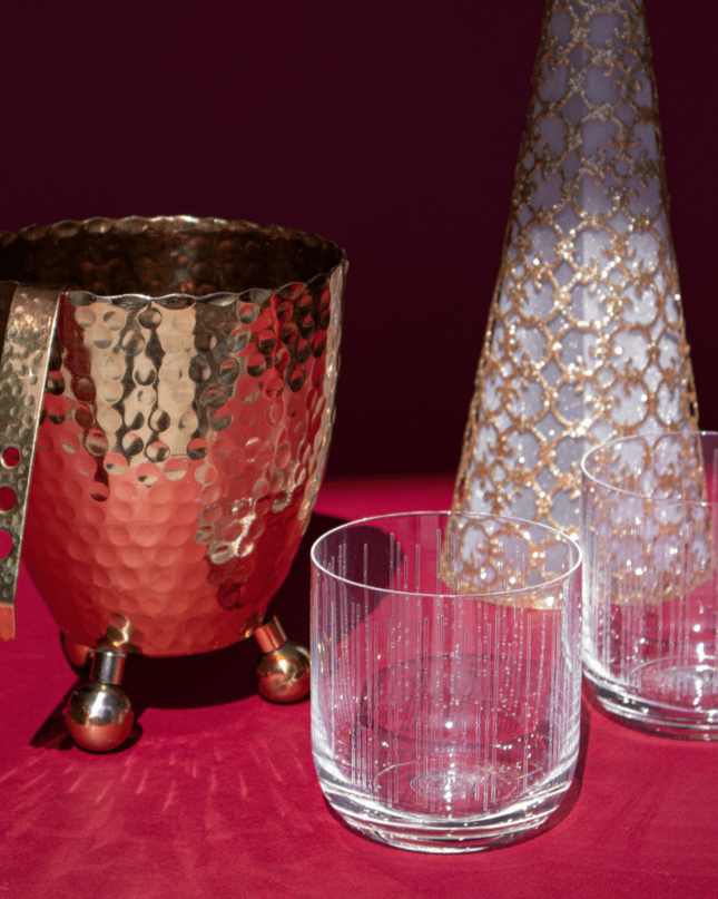 Decorative glassware including a hammered gold-toned ice bucket with tongs, gold lace Christmas tree, and clear crystal whiskey glasses on a red surface.