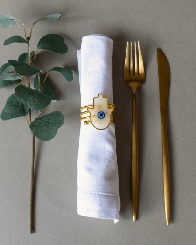Gold fork and knife with a white napkin ring featuring a hamsa design on a gray background