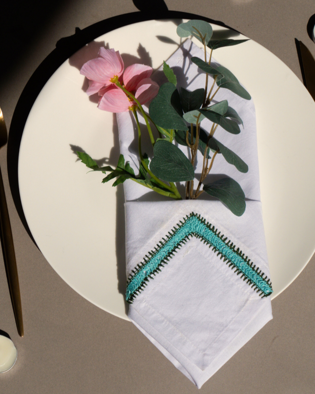 Top-down view of a table setting with a floral arrangement, hand-embroidered napkin, candles, and cutlery on a white surface.