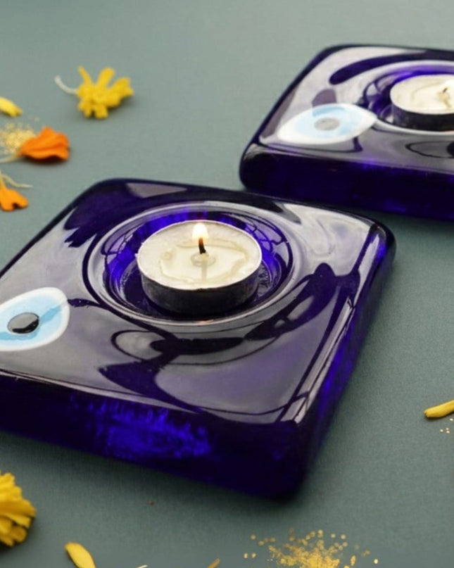 Two blue evil eye glass candle holders with candles on a dark surface with scattered flowers.