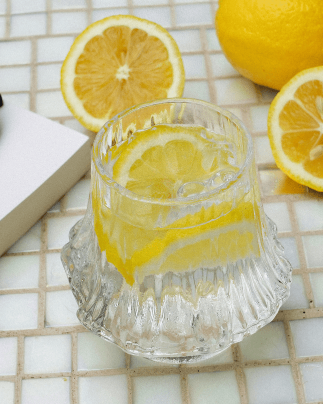 Glass of lemon water with lemons on a tiled surface near a pool.