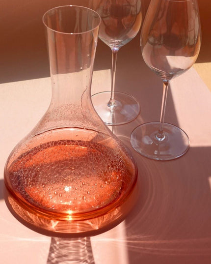 Clear glass decanter with pinkish liquid on a light surface, with two empty wine glasses in the background.