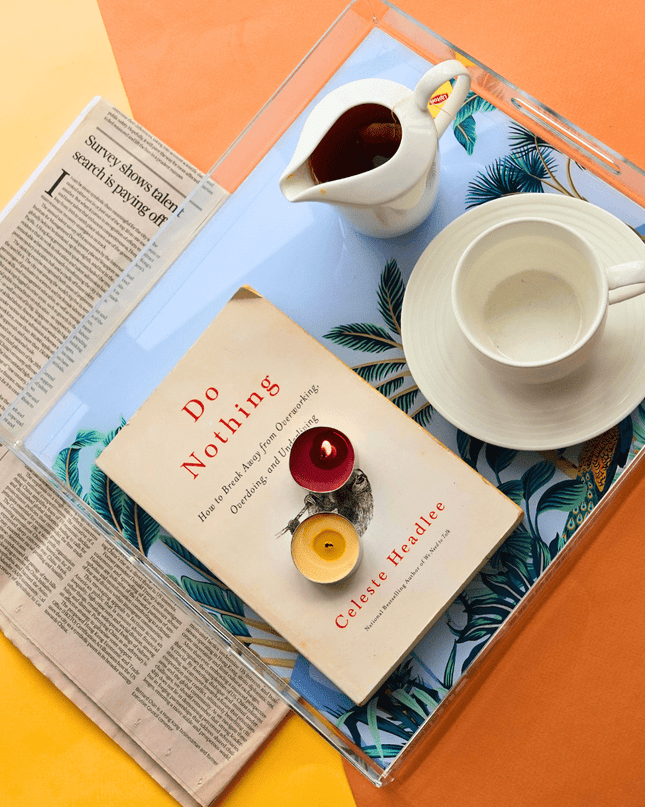Book titled 'Do Nothing' by Celeste Headlee on an elephant with palm trees acrylic tray with a cup, saucer, and candle.