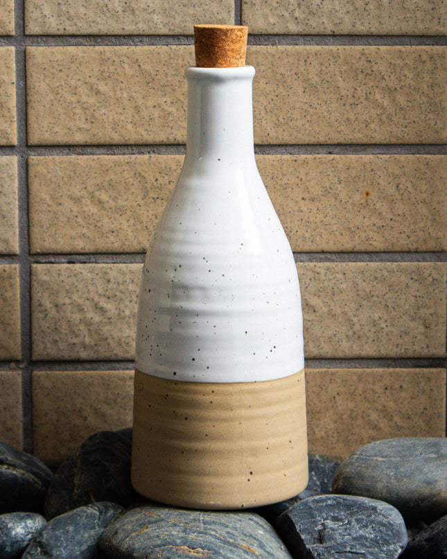 White ceramic bottle with a cork stopper on pebbles against a tiled wall.