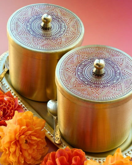 Two ornate gold brass containers with intricate designs on a pink background with orange flowers.