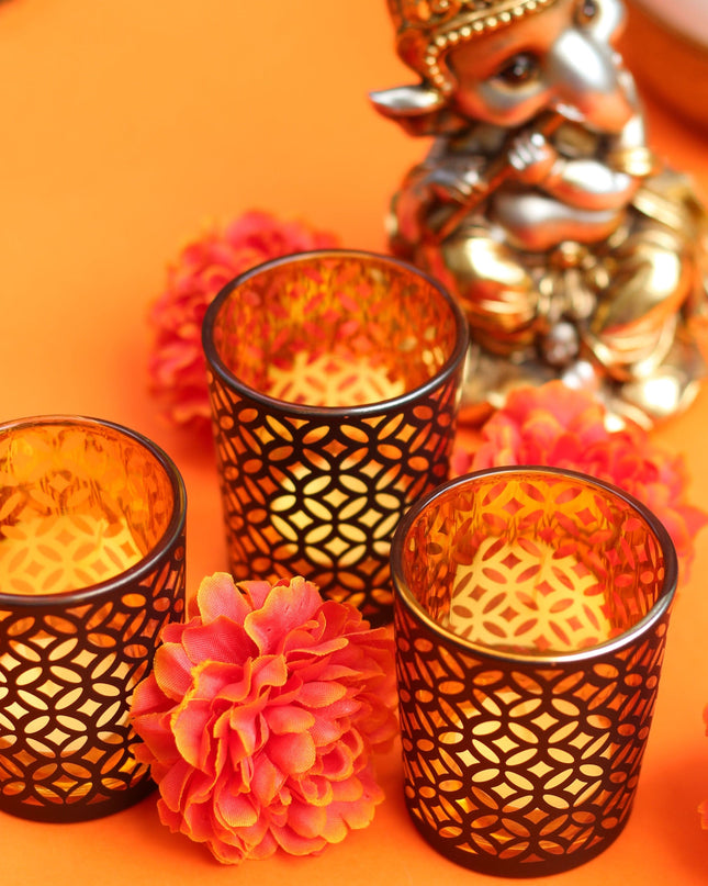 Decorative setup with three ornate black glass candle holders, a small statue of Ganesh, and pink flowers on an orange background.