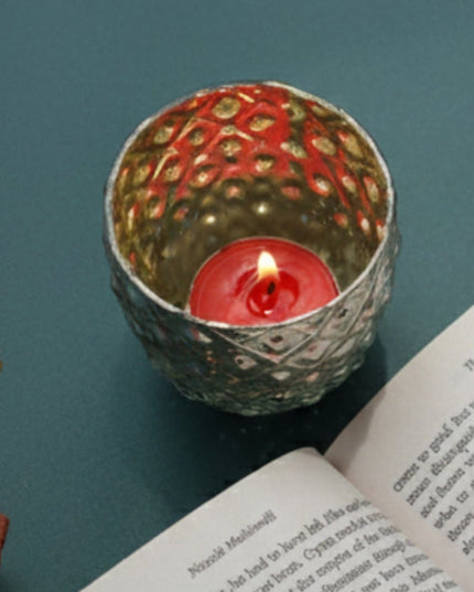 Decorative silver candle holder with a lit red candle on a teal surface next to an open book.