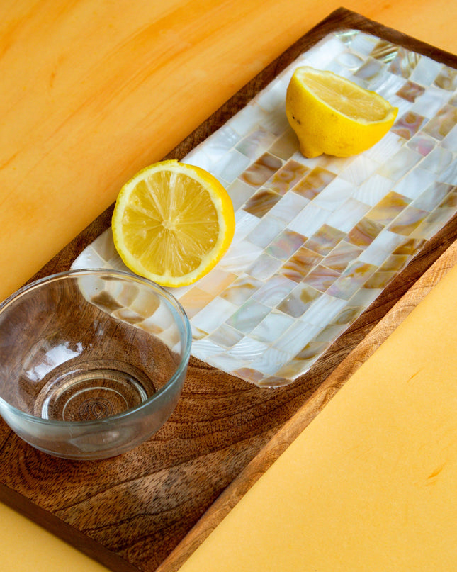 Two lemons and a glass bowl on a decorative mother of pearl and wood tray on a yellow surface.