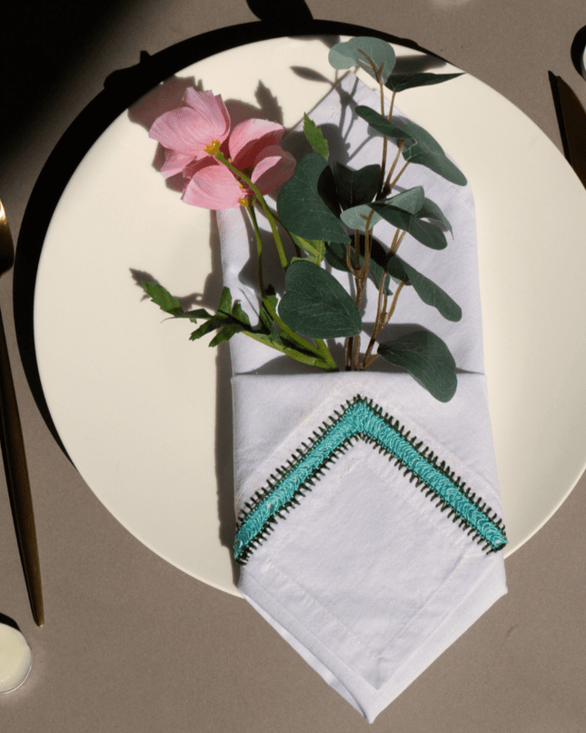 Top-down view of a table setting with a floral arrangement, hand-embroidered napkin, candles, and cutlery on a white surface.