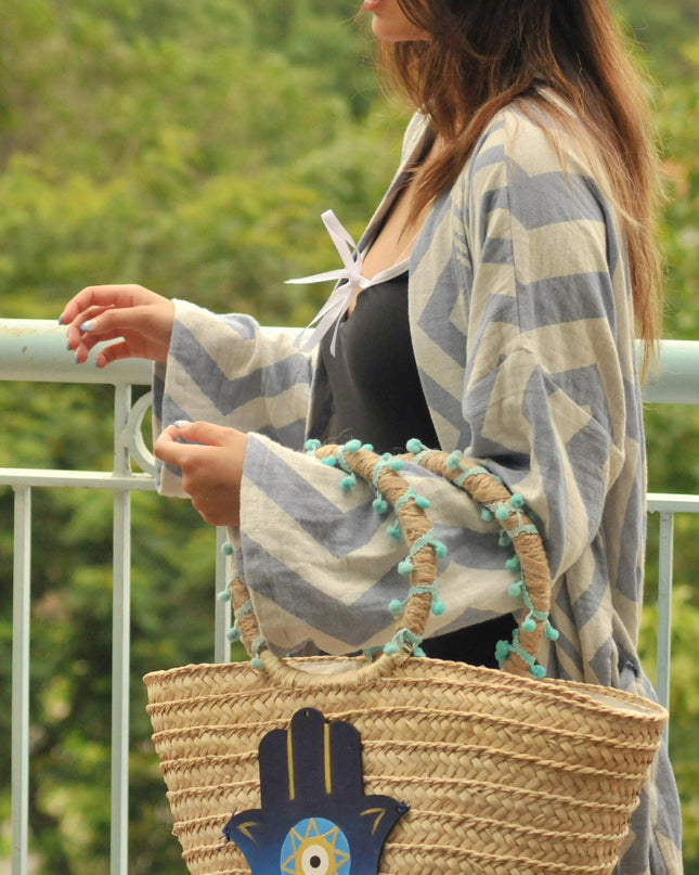 Woman wearing a straw hat and carrying a woven bag with a Hamsa hand design, standing on a balcony with greenery and buildings in the background.