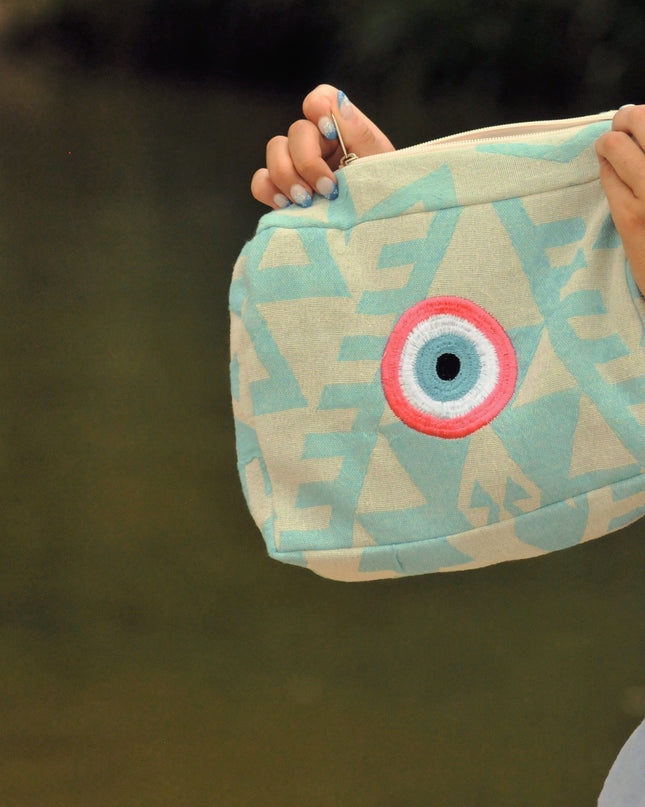 Person holding a turquoise pouch with a pink evil eye design against a blurred natural background