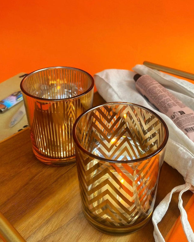 Two glass candle holders with chevron and striped patterns on a wooden surface against an orange background.