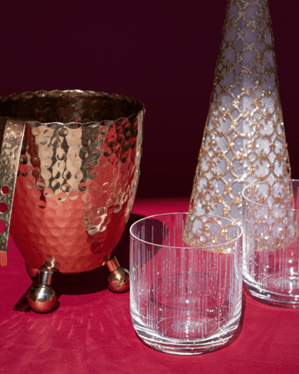 Decorative glassware including a hammered gold-toned ice bucket with tongs, gold lace Christmas tree, and clear crystal whiskey glasses on a red surface.