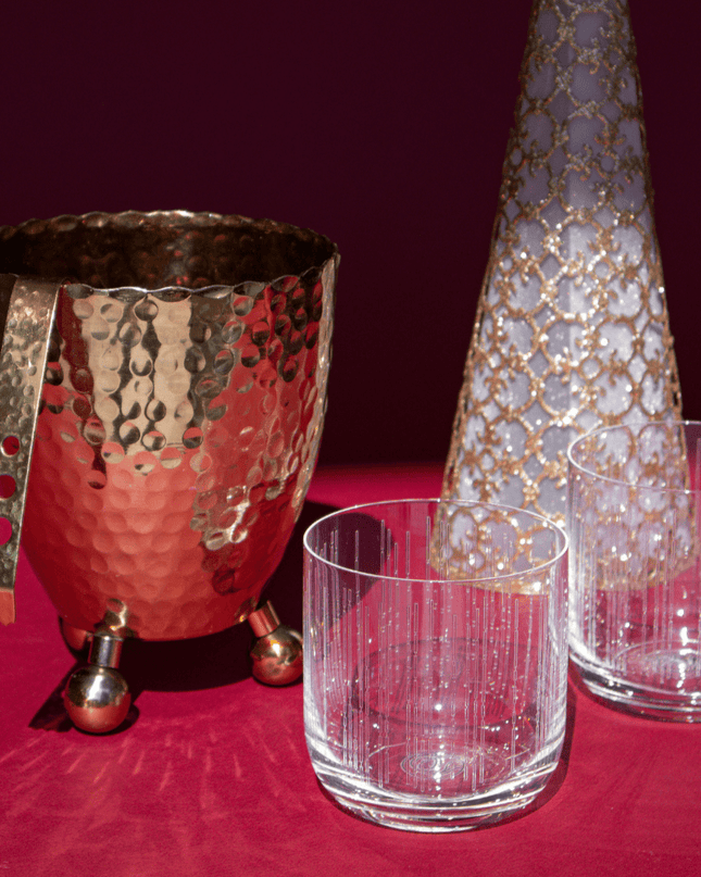Decorative glassware including a hammered gold-toned ice bucket with tongs, gold lace Christmas tree, and clear crystal whiskey glasses on a red surface.
