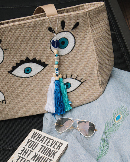 Beige tote bag with evil eye design, book, sunglasses, and towel on a dark surface