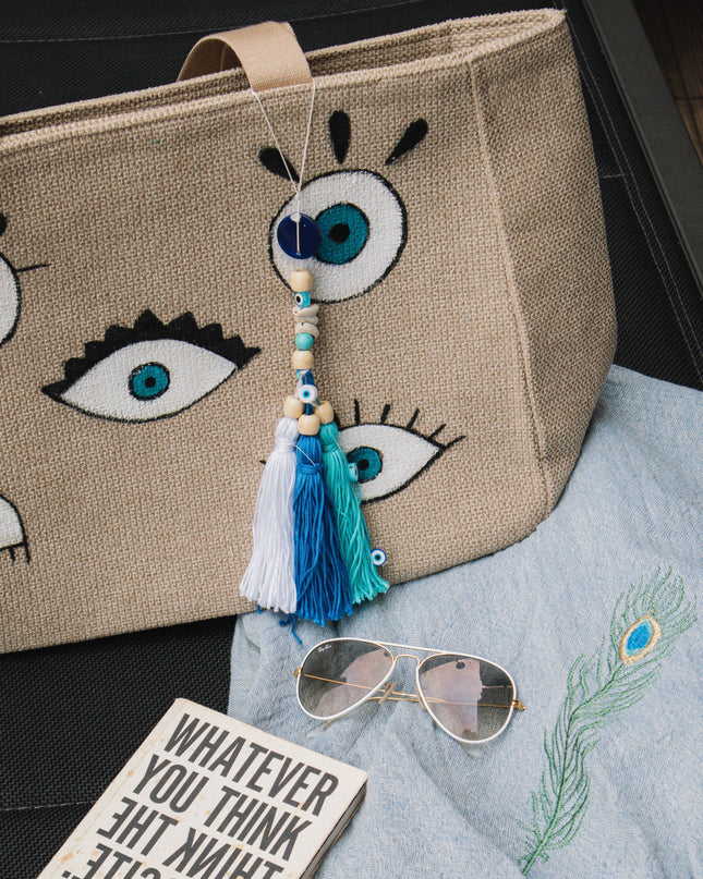 Beige tote bag with evil eye design, book, sunglasses, and towel on a dark surface