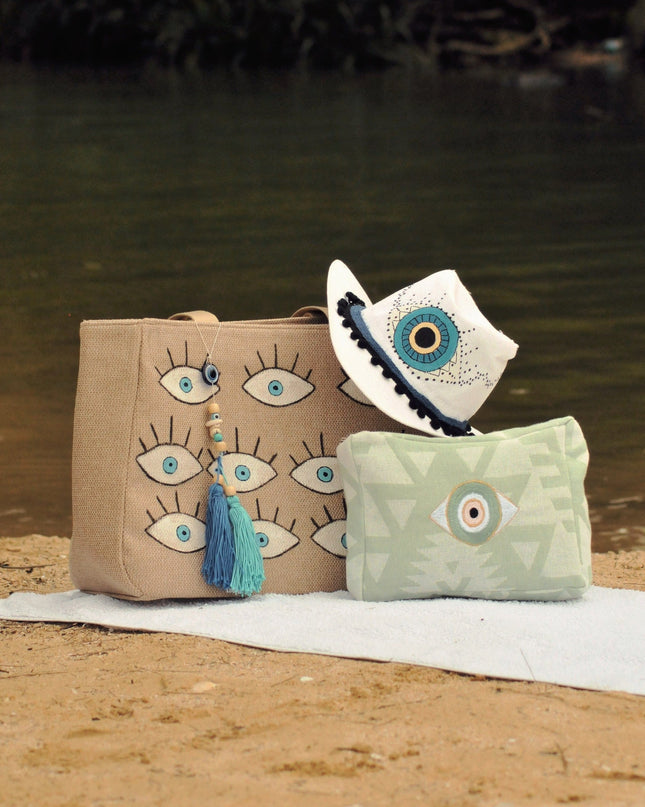 one canvas beach bag, embroidered green toiletry bag and beach hat with evil eye designs on a sandy surface near water