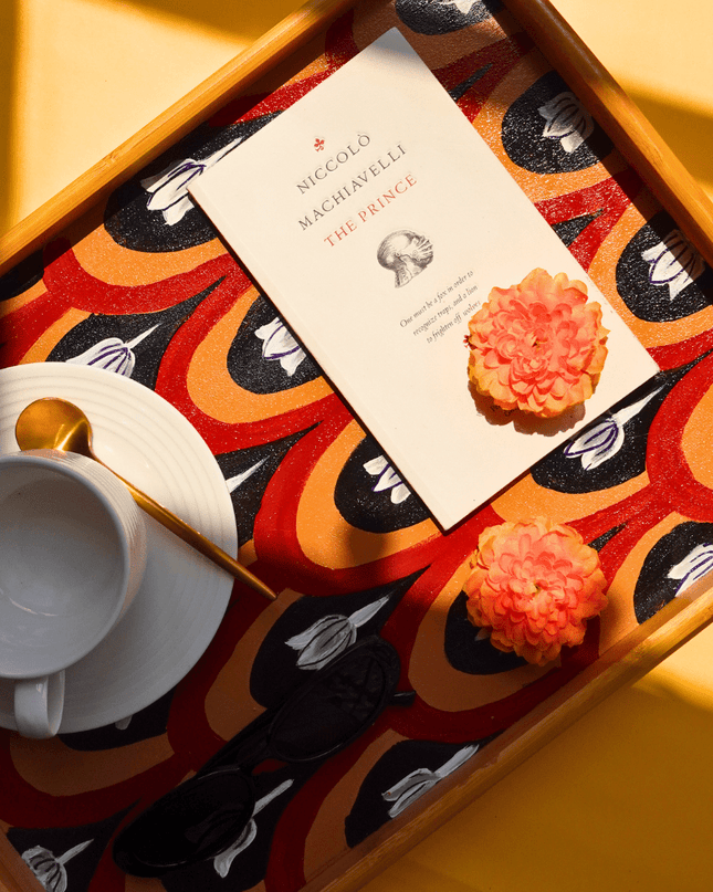 Book titled 'Niccolo Machiavelli The Prince' on a decorative hand-painted floral tray with cookies and a cup.