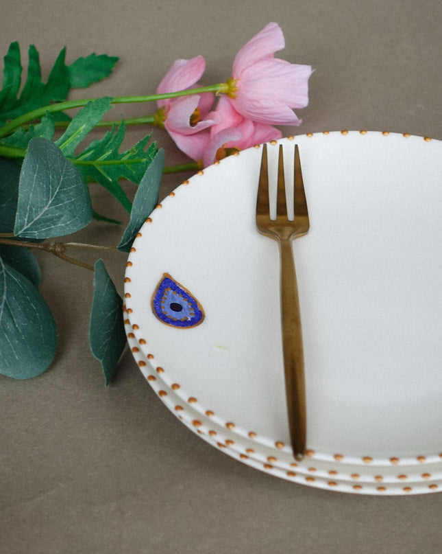 White plate with gold rim, gold fork, and decorative evil eye design on a brown background with flowers and leaves.