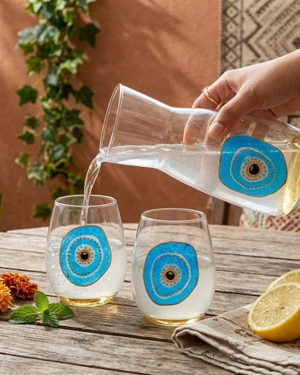 Aliya Water Carafe pouring water into two hand-painted evil eye stemless glasses on a wooden table
