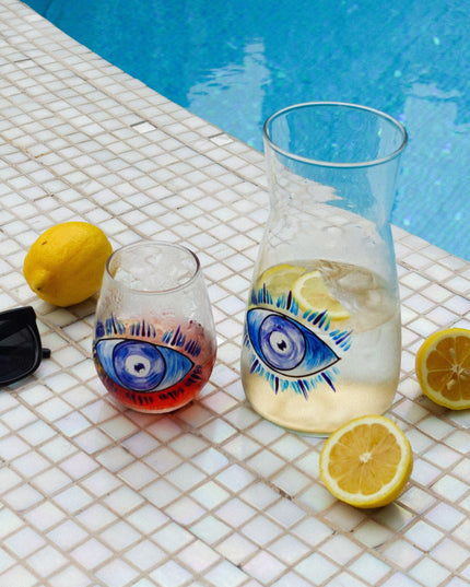 one glass and one carafe with evil eye designs and lemons on a tiled poolside surface.
