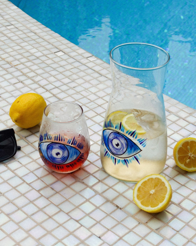 one glass and one carafe with evil eye designs and lemons on a tiled poolside surface.