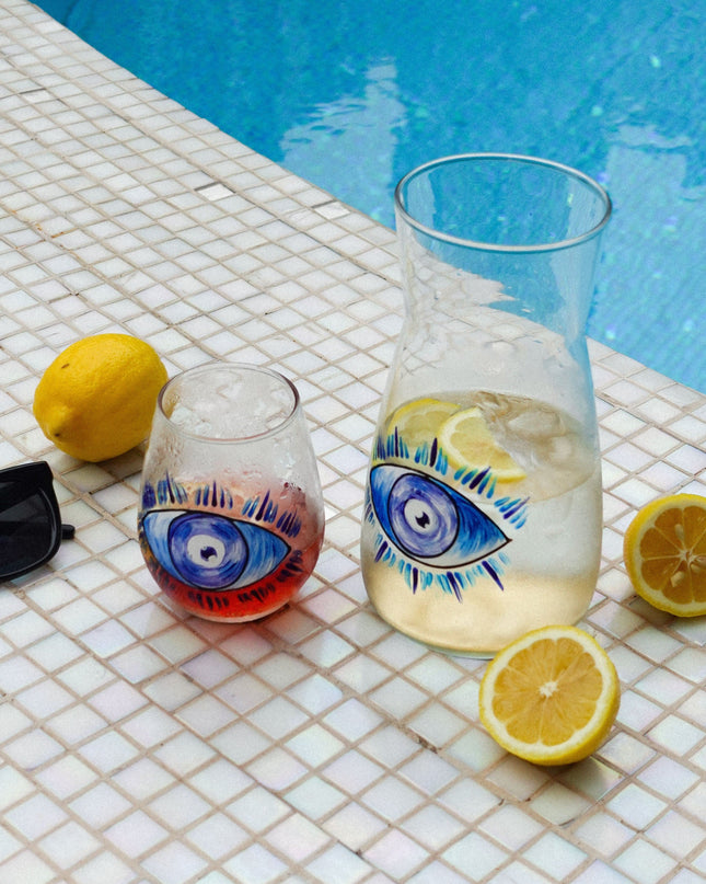 one glass and one carafe with evil eye designs and lemons on a tiled poolside surface.