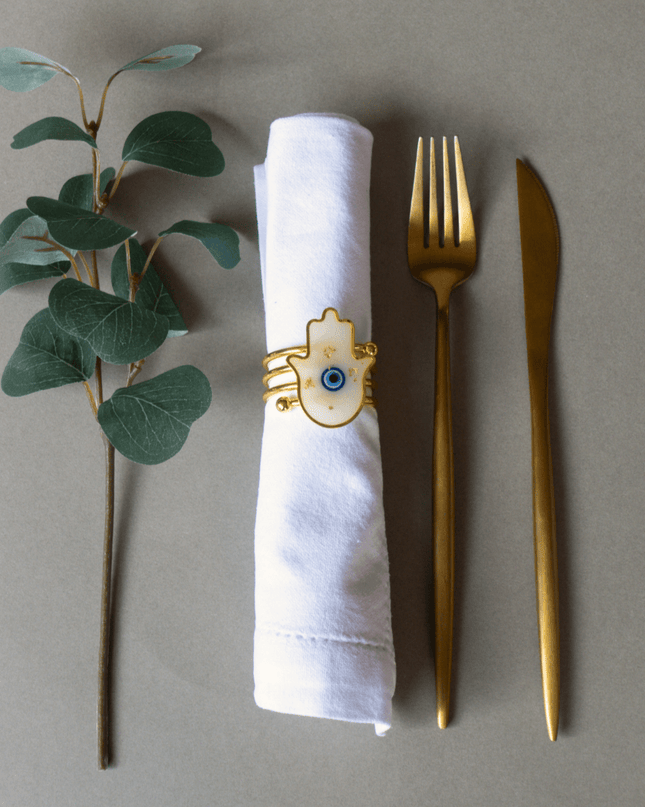 Gold fork and knife with a white napkin ring featuring a hamsa design on a gray background