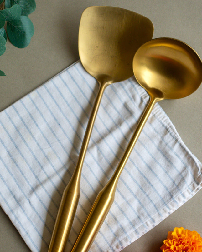 gold ladle and spatula on a white cloth with orange flowers and green leaves in the background