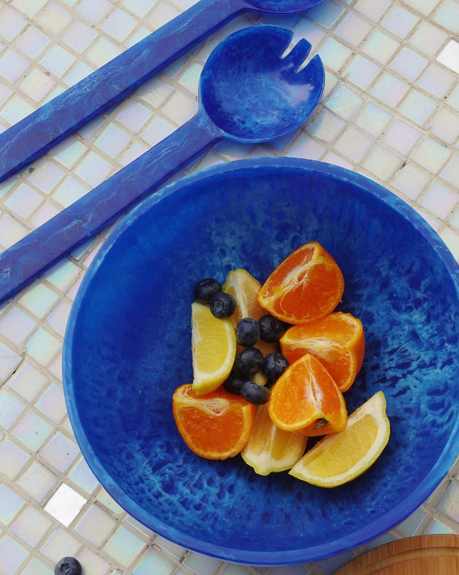 Blue resin salad bowl with fruit and salad servers on a tiled surface