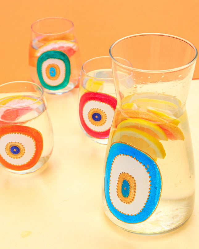 Clear glasses and water carafe with colorful evil-eye designs filled with water and lemon slices on a yellow surface.