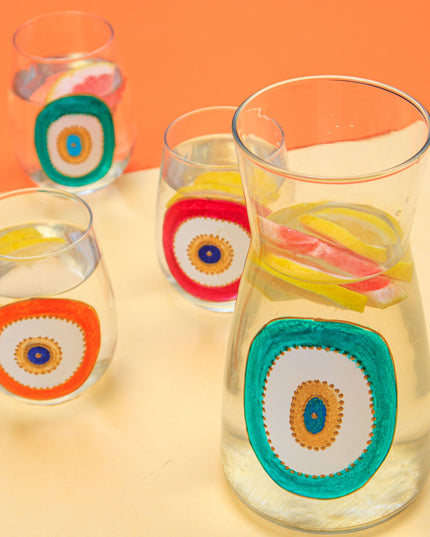 Clear glasses and water carafe with colorful evil-eye designs on a yellow and orange background