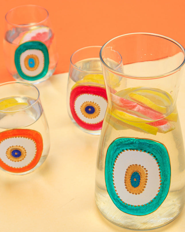 Clear glasses and water carafe with colorful evil-eye designs on a yellow and orange background